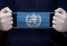 Ebola Jumps Borders – WHO on Alert Gloved hands holding a blue face mask with a medical emblem