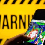 Hand holds smartphone showing cyclone radar map against warning sign