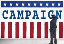 El-Sayed Undercover Video – Taking Overseas Money? Man in suit writing on large campaign banner with stars and stripes