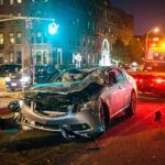 Fiery Red-Light Smash Ends In Murder Wrecked silver car on city street at night with tow truck nearby