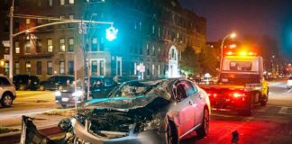 Wrecked silver car on city street at night with tow truck nearby